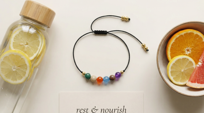 Colorful beaded bracelet on a white surface with a glass bottle, lemons, and a bowl of fruit.