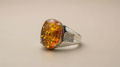 Luxury-style product photo of an oval amber cabochon ring on a reflective silver-tone setting with diamond-like accent panel.